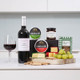 Red Wine & Cheese Gift Box. A wooden serving board displays various cheeses, crackers, and green grapes. In the background, there are two rounds of Carrigaline cheese and a jar of chutney. A bottle of red wine and a glass filled with wine are placed beside the board. The setting features a light-colored countertop against a soft blue wall. Red Wine & Cheese Gift Box. A wooden serving board displays various cheeses, crackers, and green grapes. In the background, there are two rounds of Carrigaline cheese and a jar of chutney. A bottle of red wine and a glass filled with wine are placed beside the board. The setting features a light-colored countertop against a soft blue wall.